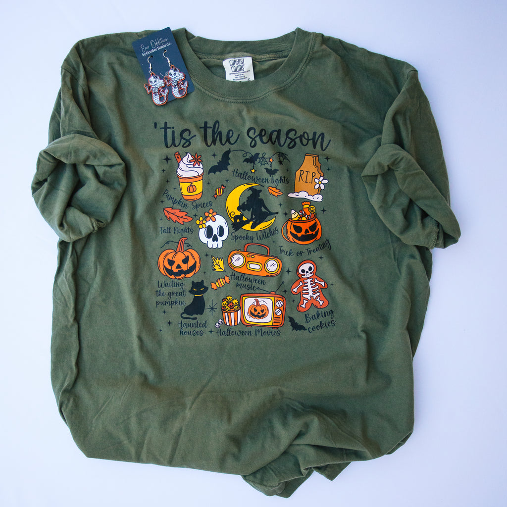An olive green long-sleeve t-shirt with a colorful Halloween graphic print on the front, laid flat on a light blue background. The graphic features 'tis the season' text along with pumpkins, bats, skulls, and candy illustrations.