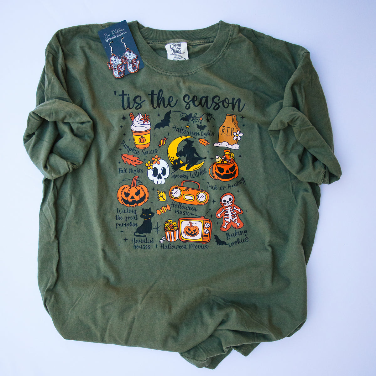 An olive green long-sleeve t-shirt with a colorful Halloween graphic print on the front, laid flat on a light blue background. The graphic features 'tis the season' text along with pumpkins, bats, skulls, and candy illustrations.