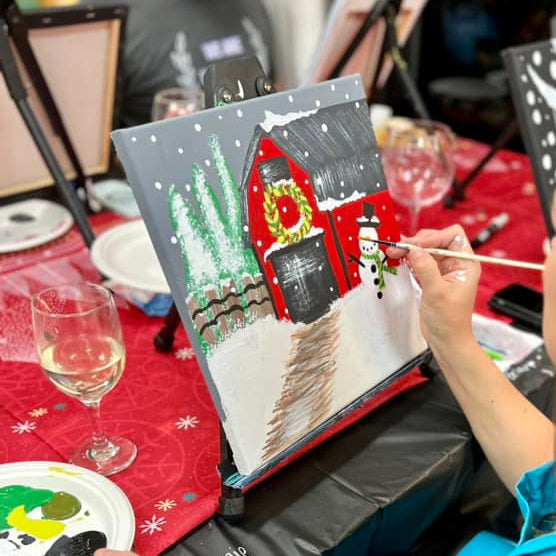 Person painting a winter scene with a red barn on a canvas held in an easel at a paint party. Paint Parties available with October House Co. in Hartville, Ohio.